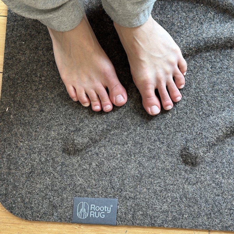 A close up of a pair of feet standing on a brown Rooty Rug that has uneven floor texture to stimulate the feet