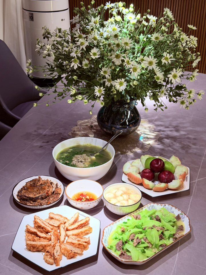 Một bữa cơm mang đậm nét truyền thống: Thịt bò xào su su, chả cá chiên giòn, cà pháo muối, canh cua, cá kho và đĩa trái cây mát lành. Không khí dịu dàng được tô điểm thêm bởi bình hoa cúc họa mi trắng tinh khôi.